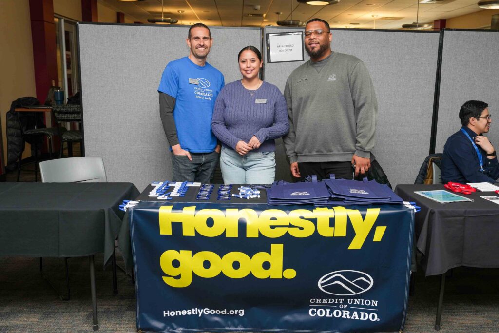Three smiling people stand behind a table adorned with a banner that reads "Honestly, good. Credit Union of Colorado.” The setting is a well-lit indoor event space.