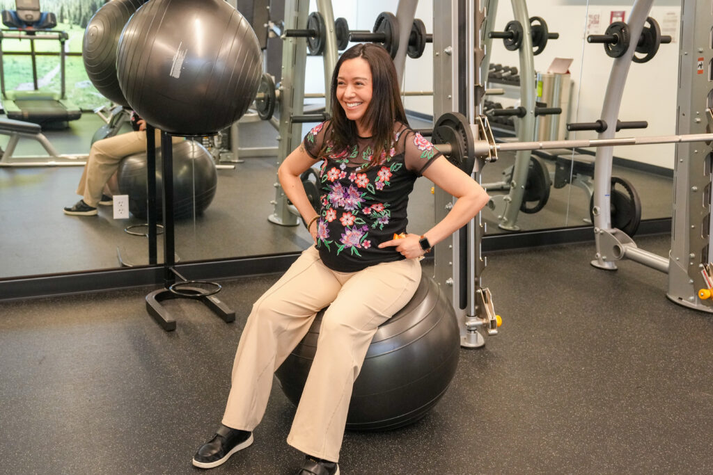 A smiling woman sits on an exercise ball in a gym. She wears a floral top and beige pants. Weightlifting equipment is visible in the background.