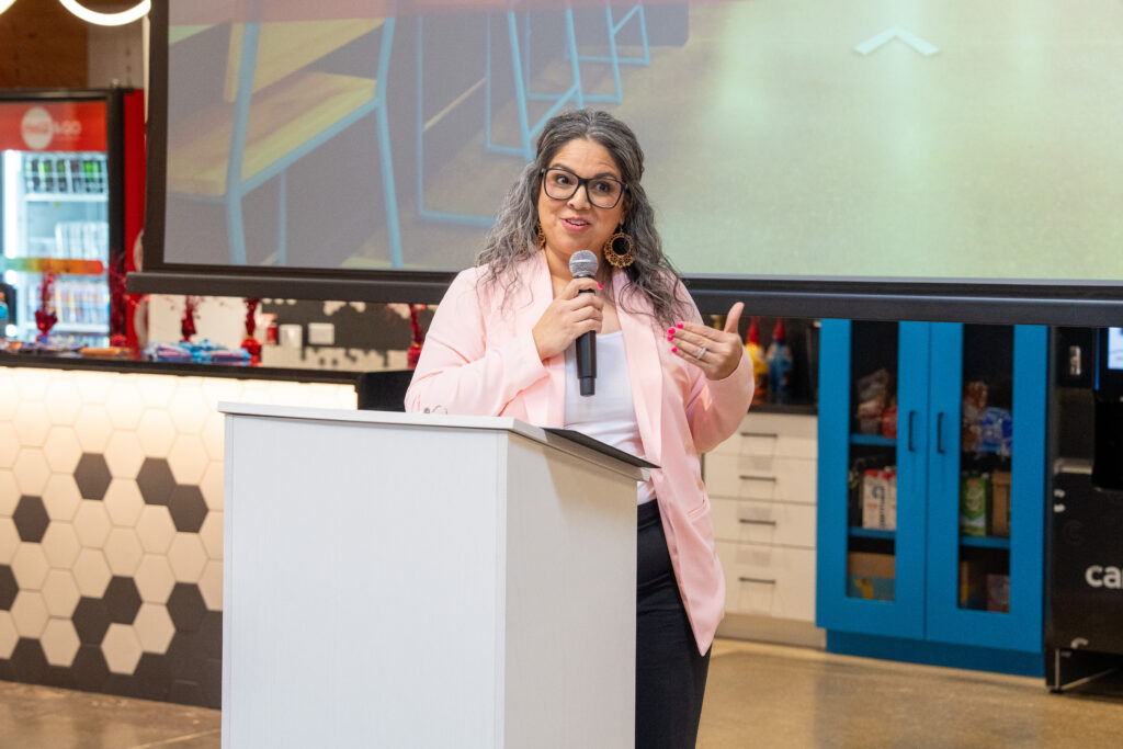 A woman with glasses and a pink sweater speaks at a podium, holding a microphone. She appears engaged and expressive in a modern setting.