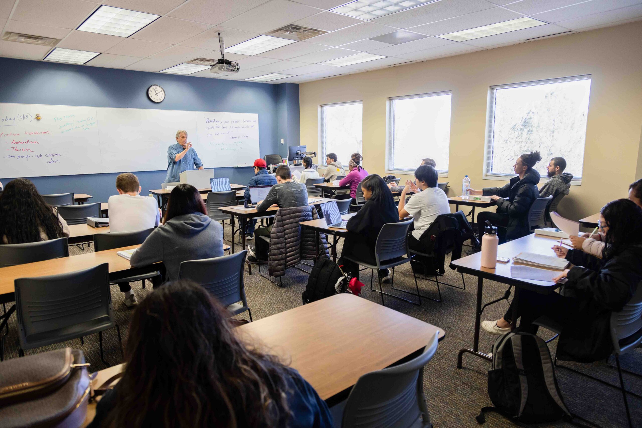 A classroom with students seated at desks, facing a teacher at the whiteboard. Bright windows illuminate the room, creating a focused learning atmosphere.