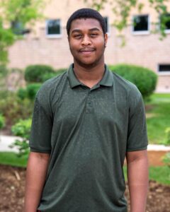 Young man in a dark green polo stands outdoors, smiling slightly. The background features lush green shrubs and a brick building, giving a serene tone.