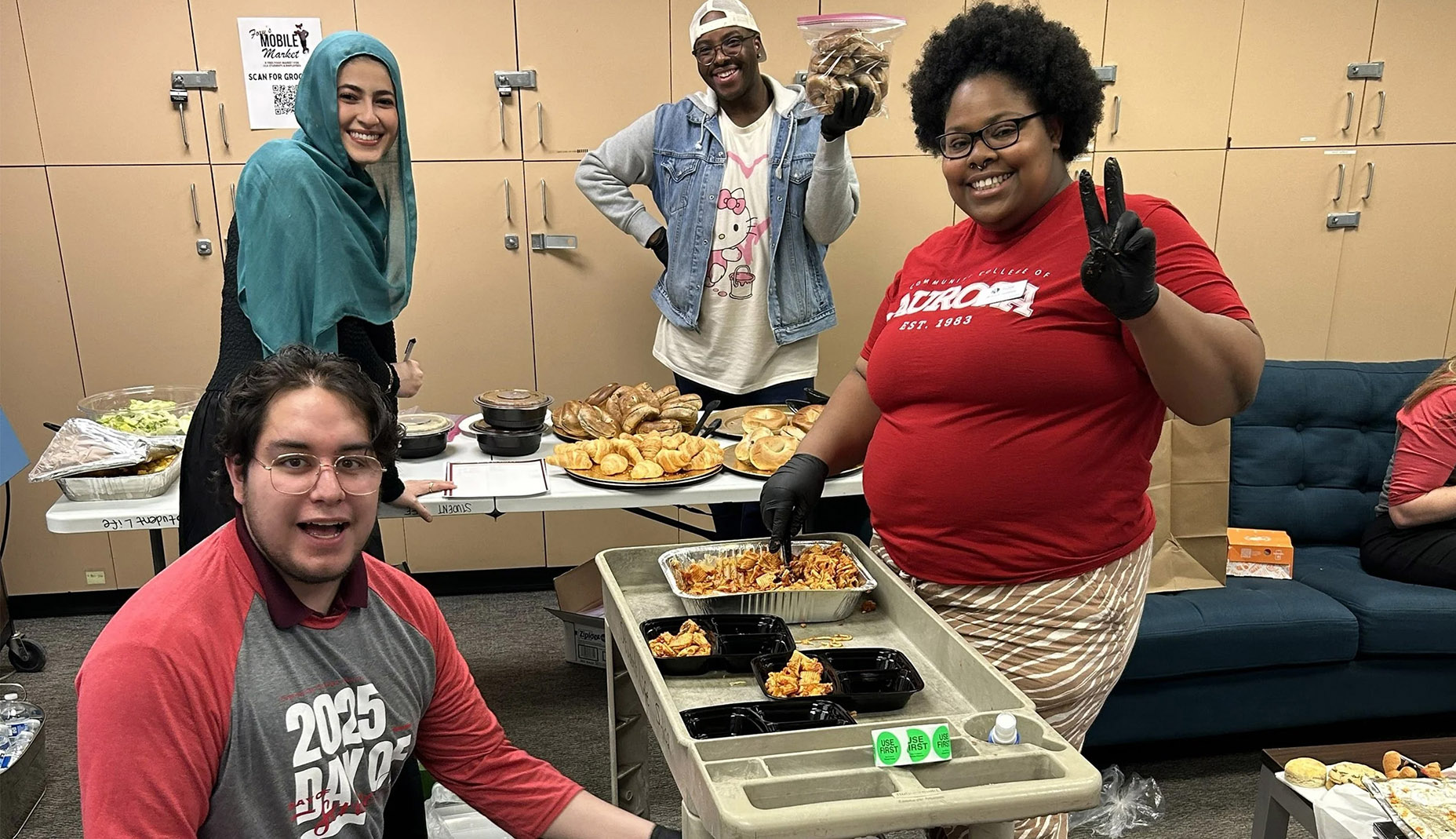 Four people smiling while distributing food in a room with cabinets. The atmosphere is lively and collaborative, with various dishes on trays and tables.