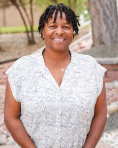 Smiling woman with short hair and patterned blouse stands outdoors, with trees and a rocky garden in the blurred background, conveying warmth.