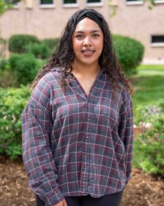 Young woman with curly hair and a headband, wearing a plaid shirt, stands outdoors by green bushes and a brick building. She smiles softly.