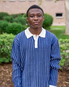 A person wearing a blue and white striped sweater stands outside, surrounded by greenery and bushes. The expression is calm, with a soft background.