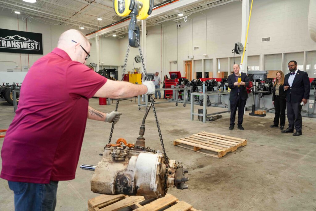Photo of four people standing in a garage setting. In the foreground, one of them maneuvers an engine as it is suspended by chains from a crane. In the background, one person operates the crane and the others observe.