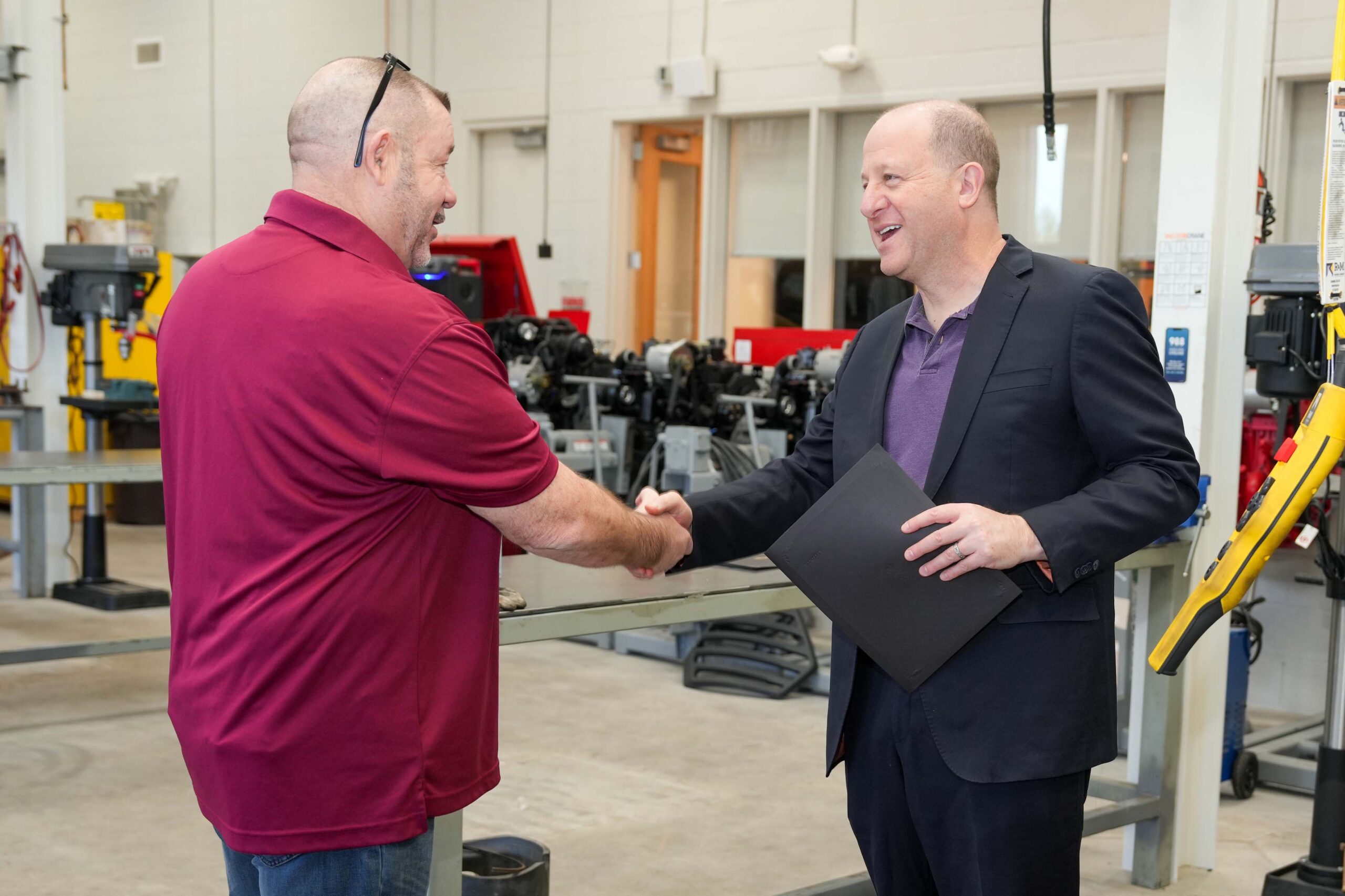 Two men shake hands in a technical training lab, with machinery and equipment visible in the background, as one holds a folder.