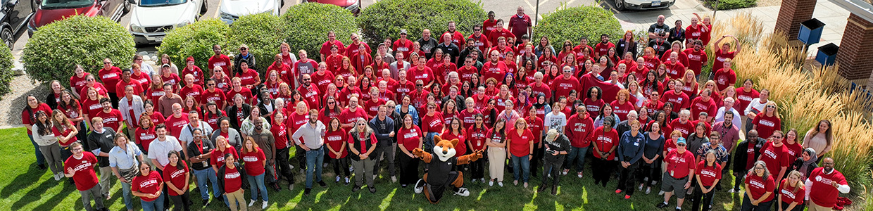 A large group of people wearing red shirts stand in a grassy area posing for a photo. There's a cheerful atmosphere, with greenery and parked cars in the background.