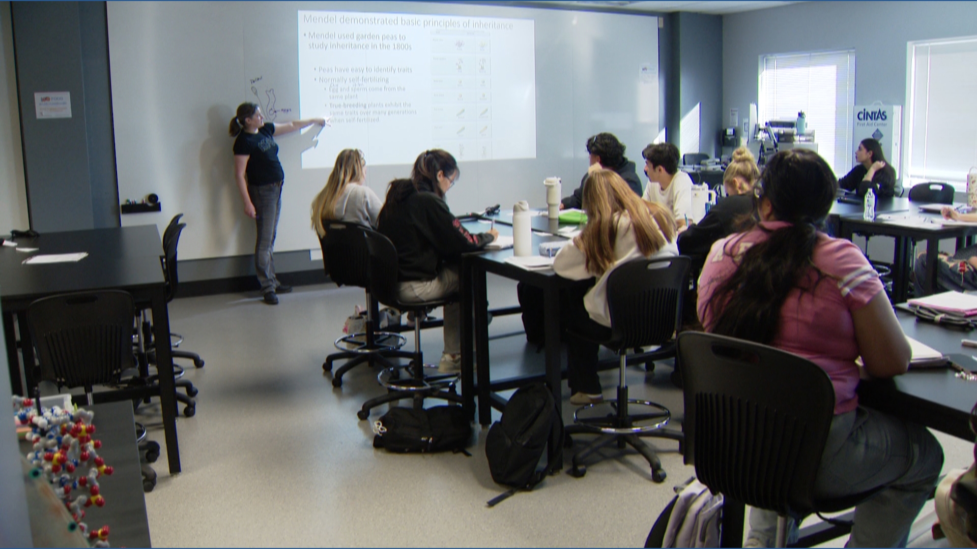 A classroom with students seated at tables, attentively watching a teacher pointing at a projected slide on a whiteboard. The atmosphere is focused and educational.