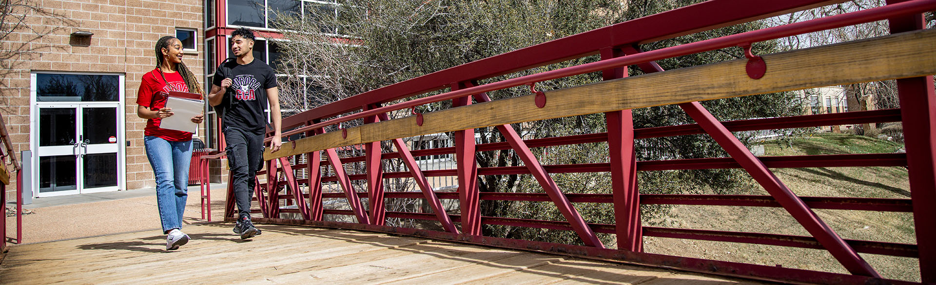 A woman and a man walk on a red bridge outside, under a clear sky. She carries a notebook and both are smiling, suggesting a friendly conversation.