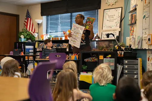 A teacher reads aloud to attentive young students in a colorful classroom. The atmosphere is lively and engaging, with educational posters and an American flag displayed.