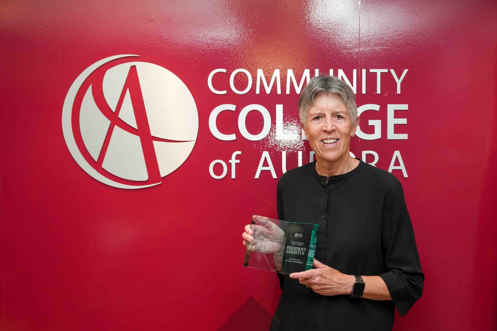 A person in a black shirt stands smiling in front of a red background with the Community College of Aurora logo while holding a glass award that reads ‘President Emeritus Dr. Oudenhoven.’