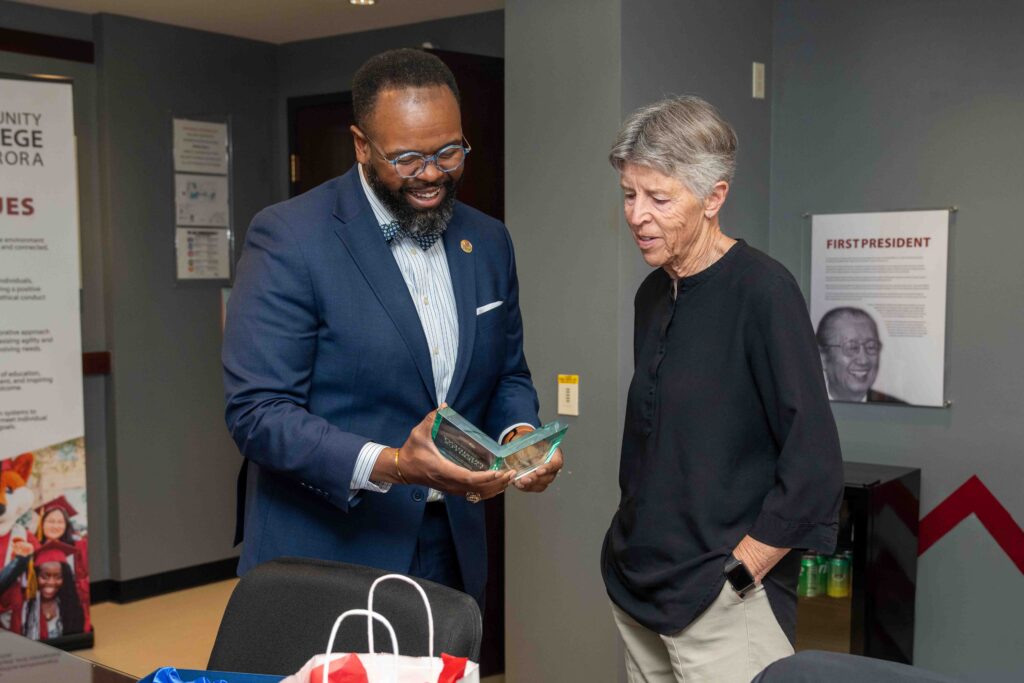 A man in a blue suit presents an award to a smiling woman in a black shirt. They stand in an office with a banner and framed photo on the wall.