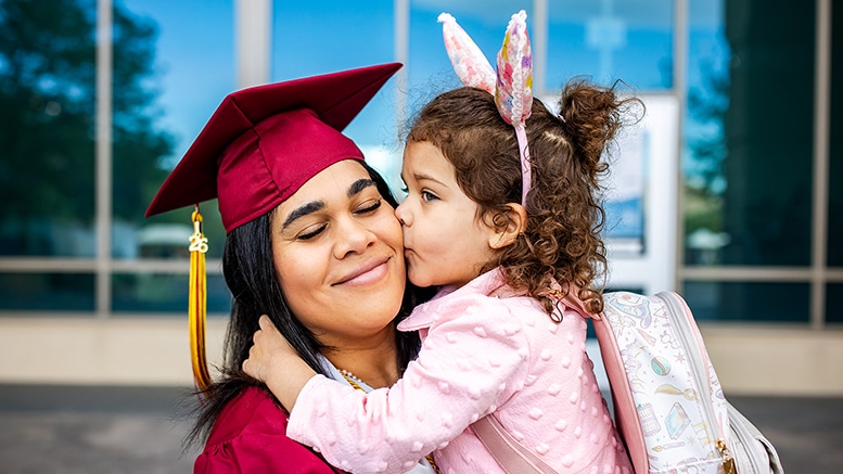 Graduation celebration: A woman in a maroon cap and gown receives a loving kiss from a young child in bunny ears, conveying joy and pride.