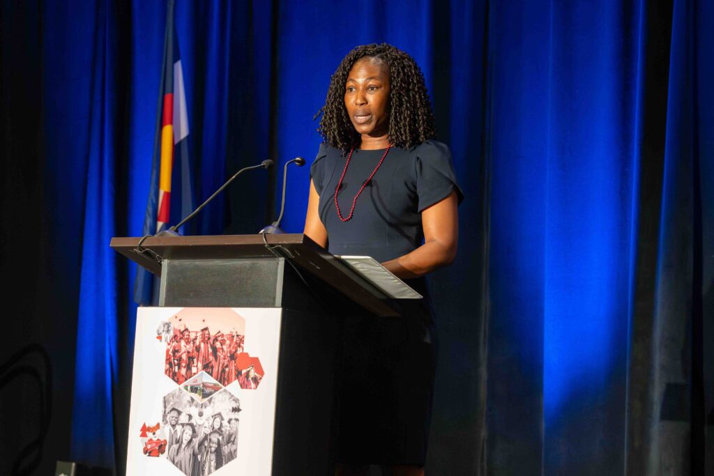 A woman stands at a podium speaking, with a blue curtain backdrop and a flag visible. She wears a navy dress and red necklace, conveying confidence.