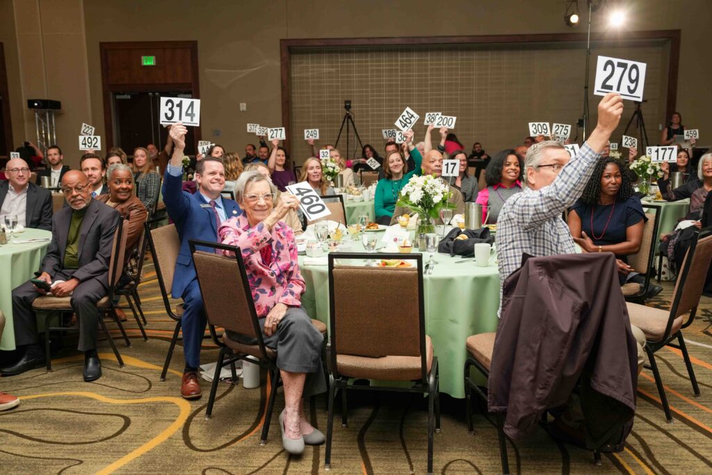 People seated at round tables in a banquet hall enthusiastically raise numbered cards for donating. The atmosphere is lively and engaging.