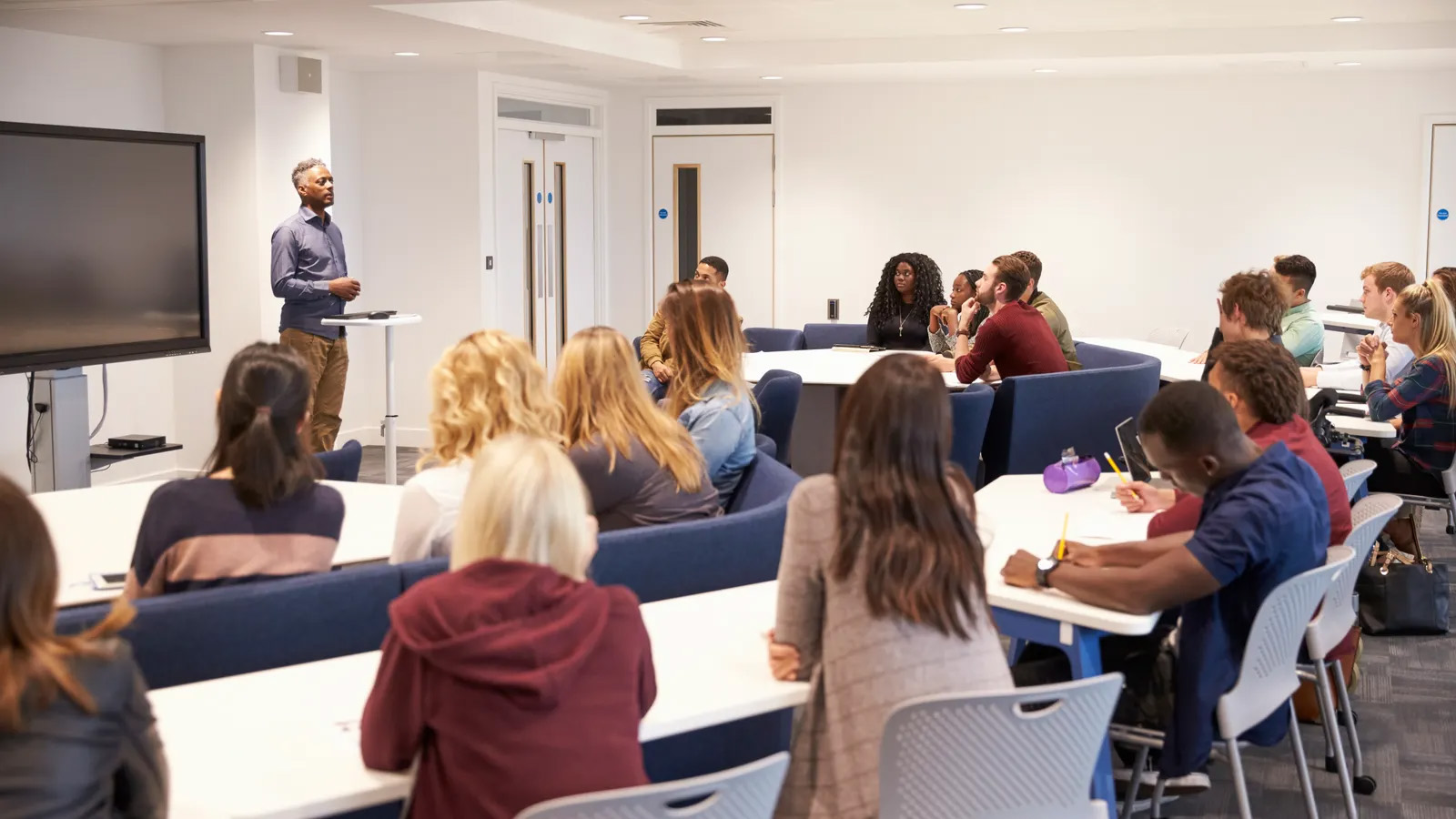 A lecturer stands at the front of a modern classroom, addressing a diverse group of attentive university students seated at tables, fostering a collaborative learning environment.