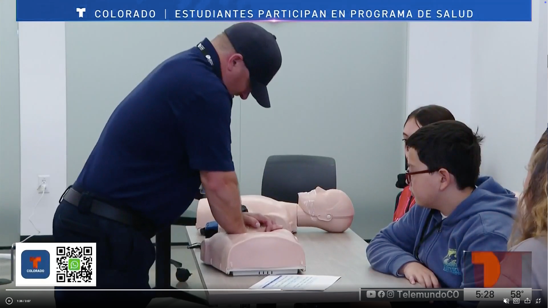 A person demonstrates CPR on a mannequin in a classroom. Students attentively watch, seated around a table, conveying an educational and focused atmosphere.