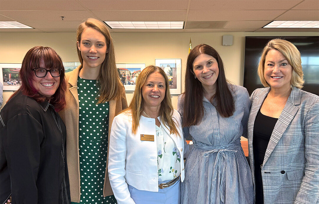 Five women in professional clothes stand together in an office setting smiling.