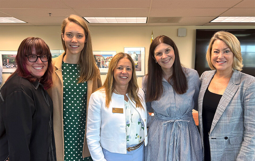 Five women in professional clothes stand together in an office setting smiling.
