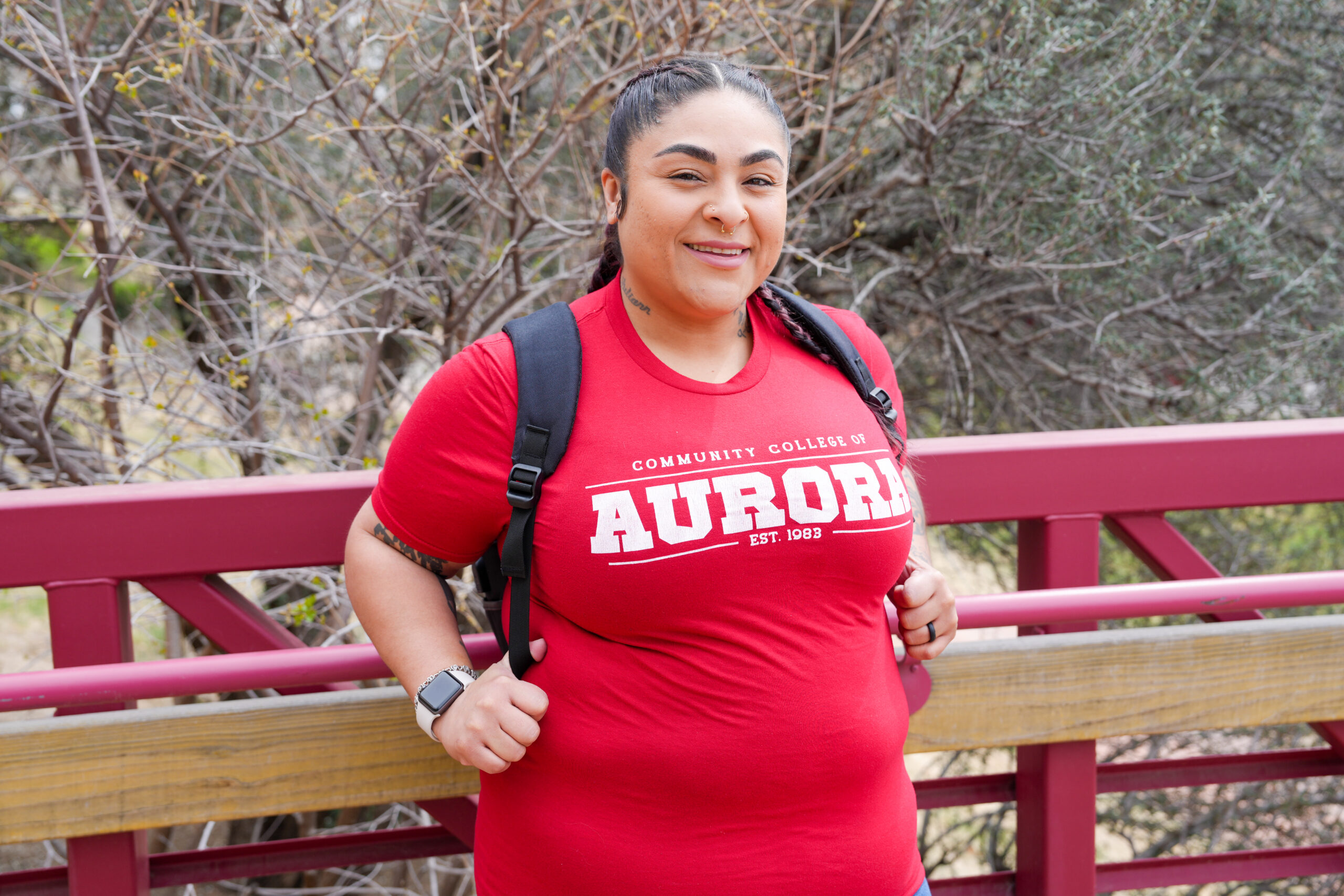 CCA student wearing a red Community College of Aurora T-shirt and backpack smiles while standing on a campus bridge outdoors.