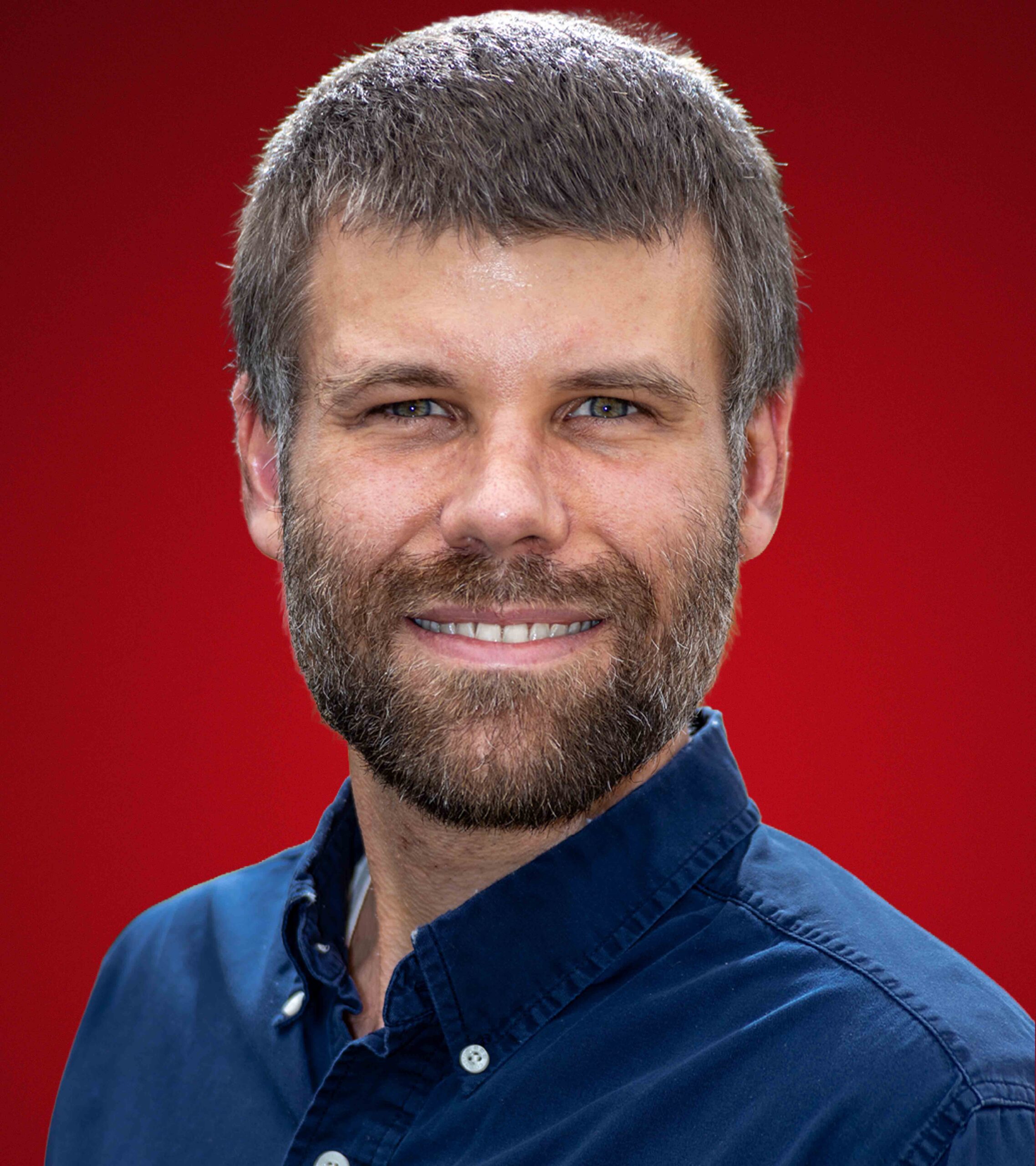 Man with short hair and beard smiling, wearing a blue button-up shirt against a vibrant red background. The mood is warm and approachable.