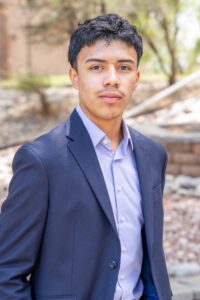 Young man in a blue blazer and light blue shirt stands outside, looking confident. The background is a sunny, blurred garden with trees.