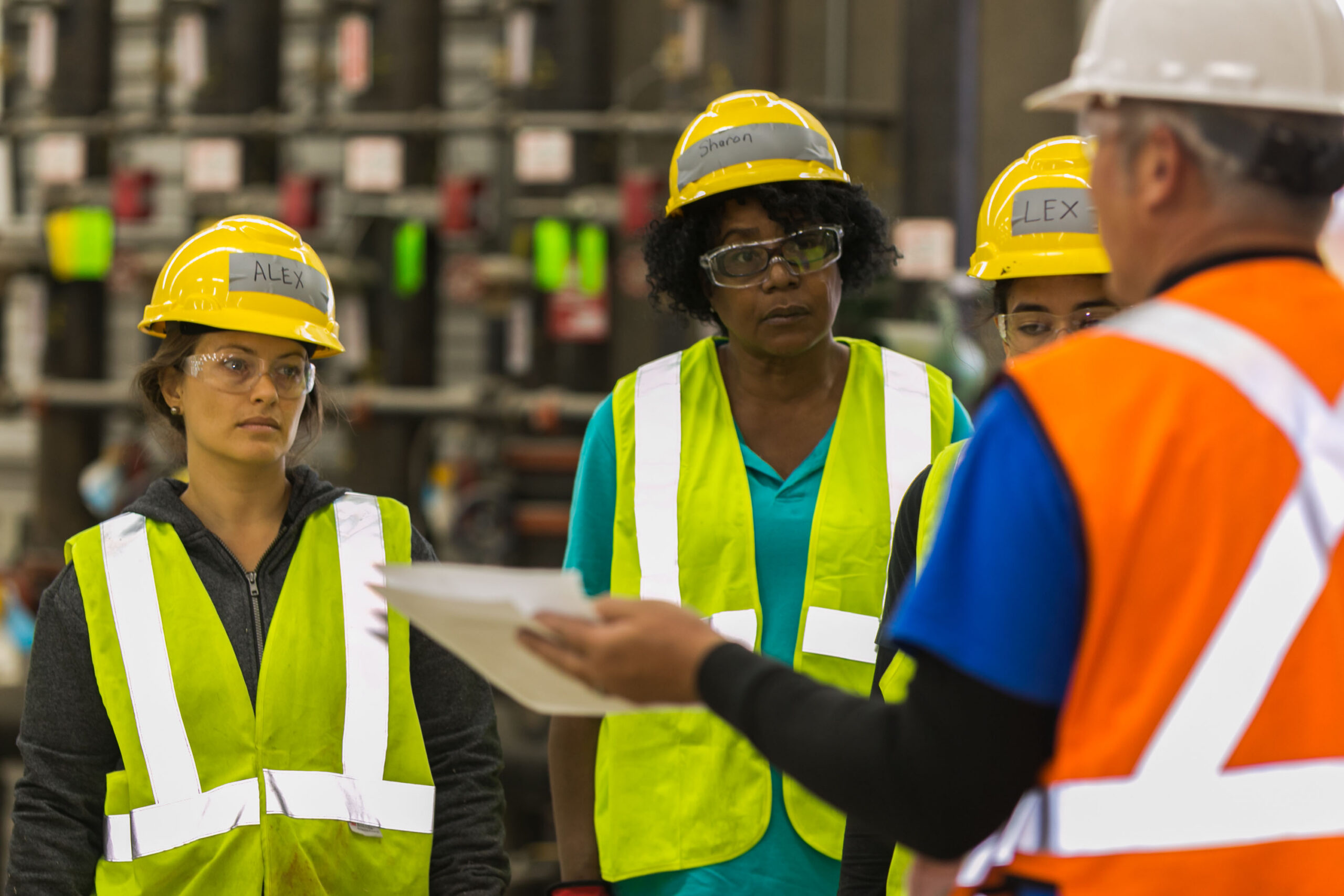 Four workers wearing safety vests and helmets listen as a supervisor holds documents during a discussion in an industrial setting.