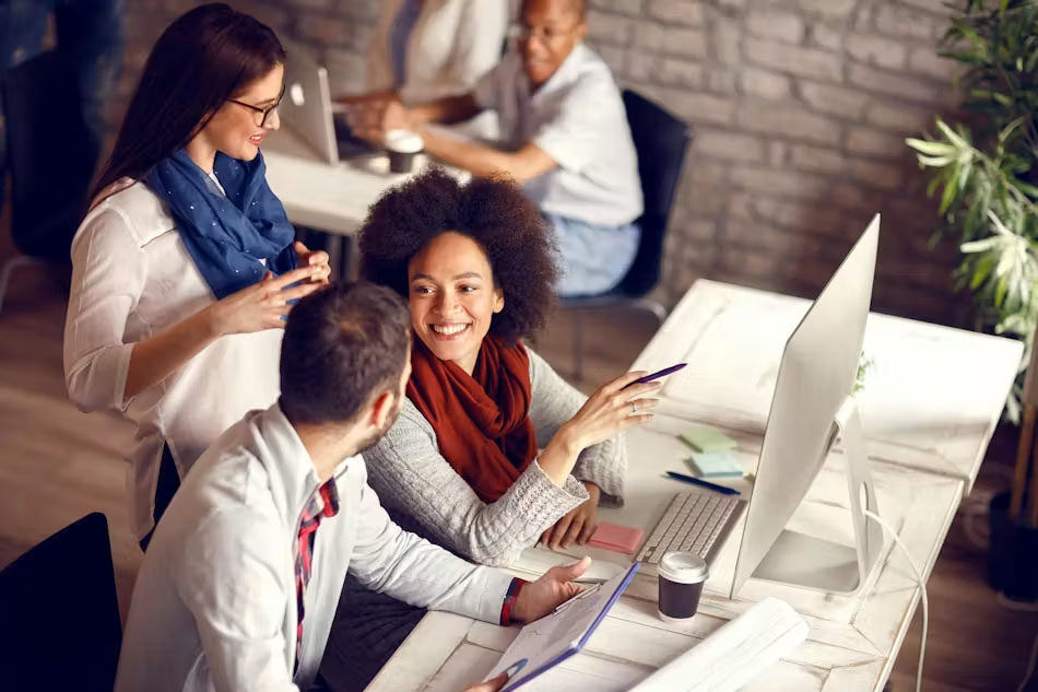 A group of four coworkers collaborates in a modern office, with one person smiling and gesturing with a pen while speaking to two colleagues at a desk with a large desktop computer, as another coworker works in the background.