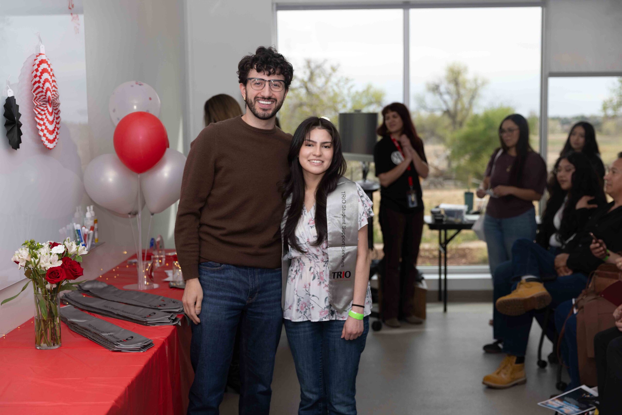Un hombre y una mujer están de pie juntos, sonriendo, en una fiesta animada con decoraciones coloridas de fondo.