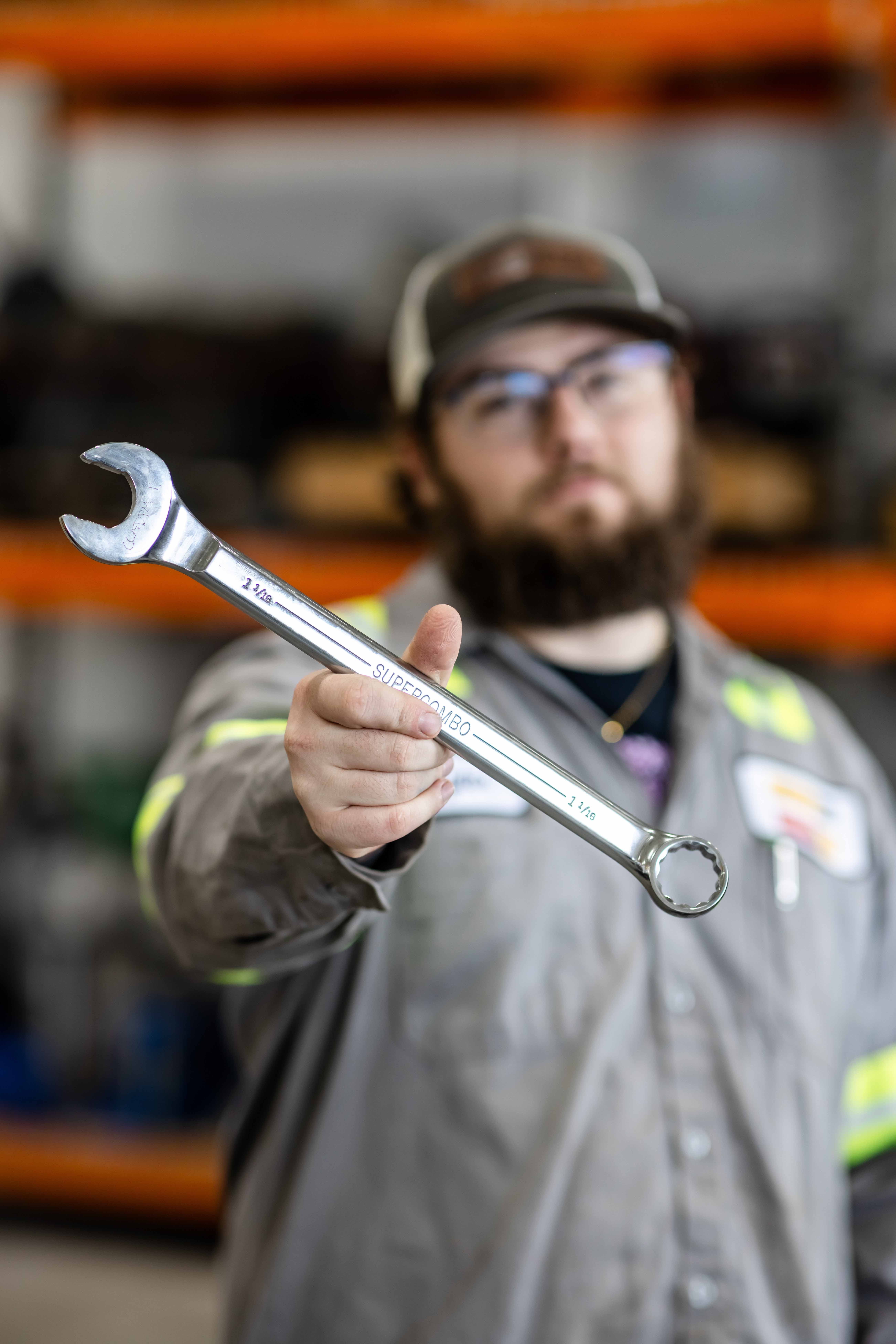 Person in work uniform extending a large combination wrench toward the camera in a workshop with industrial shelving in the background.