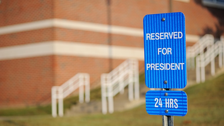 Blue sign with 'Reserved for President 24 HRS' in white text, stands near a brick building, with white railings blurred in the background.