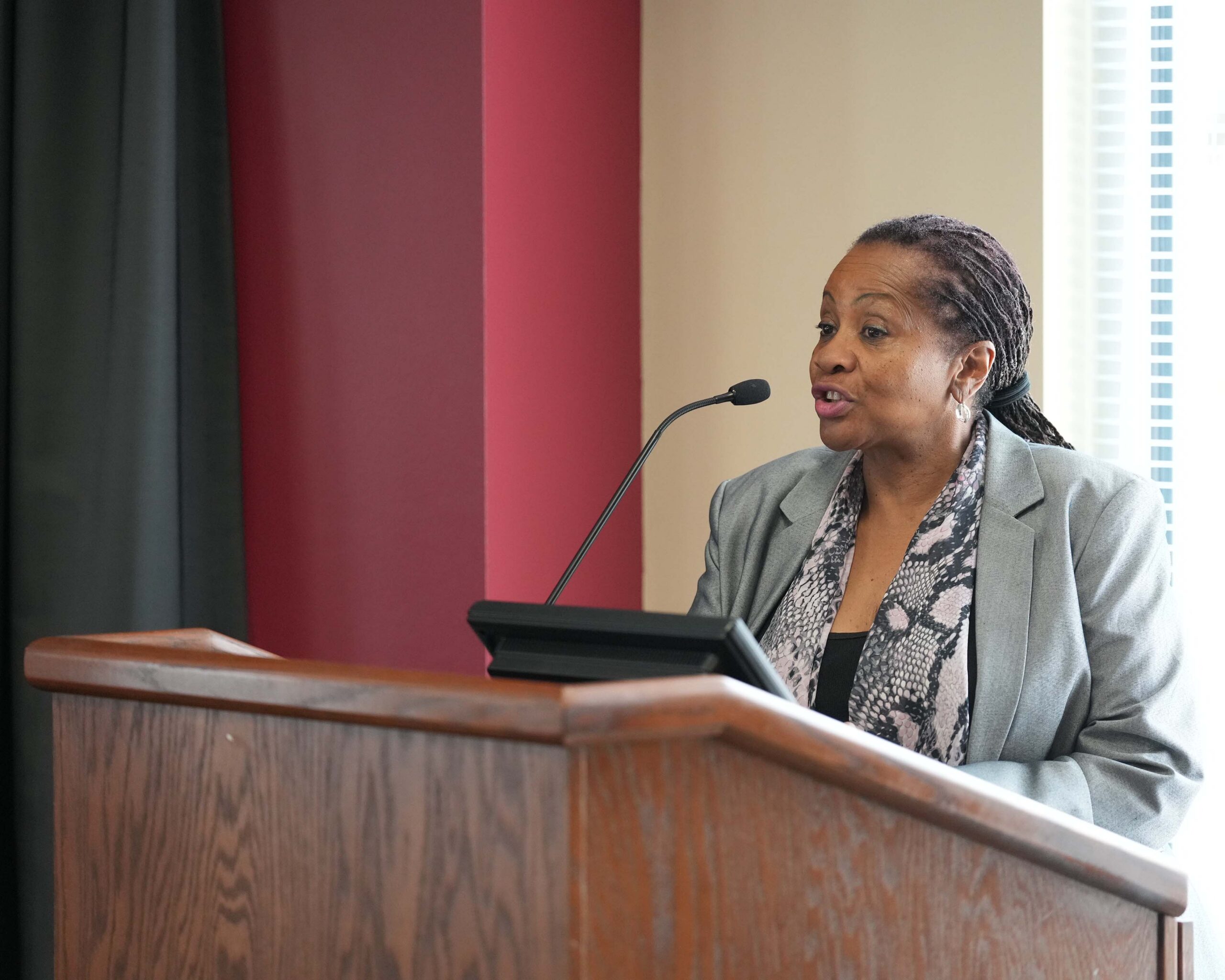 A speaker stands at a podium addressing an audience during an event, speaking into a microphone in a meeting room.