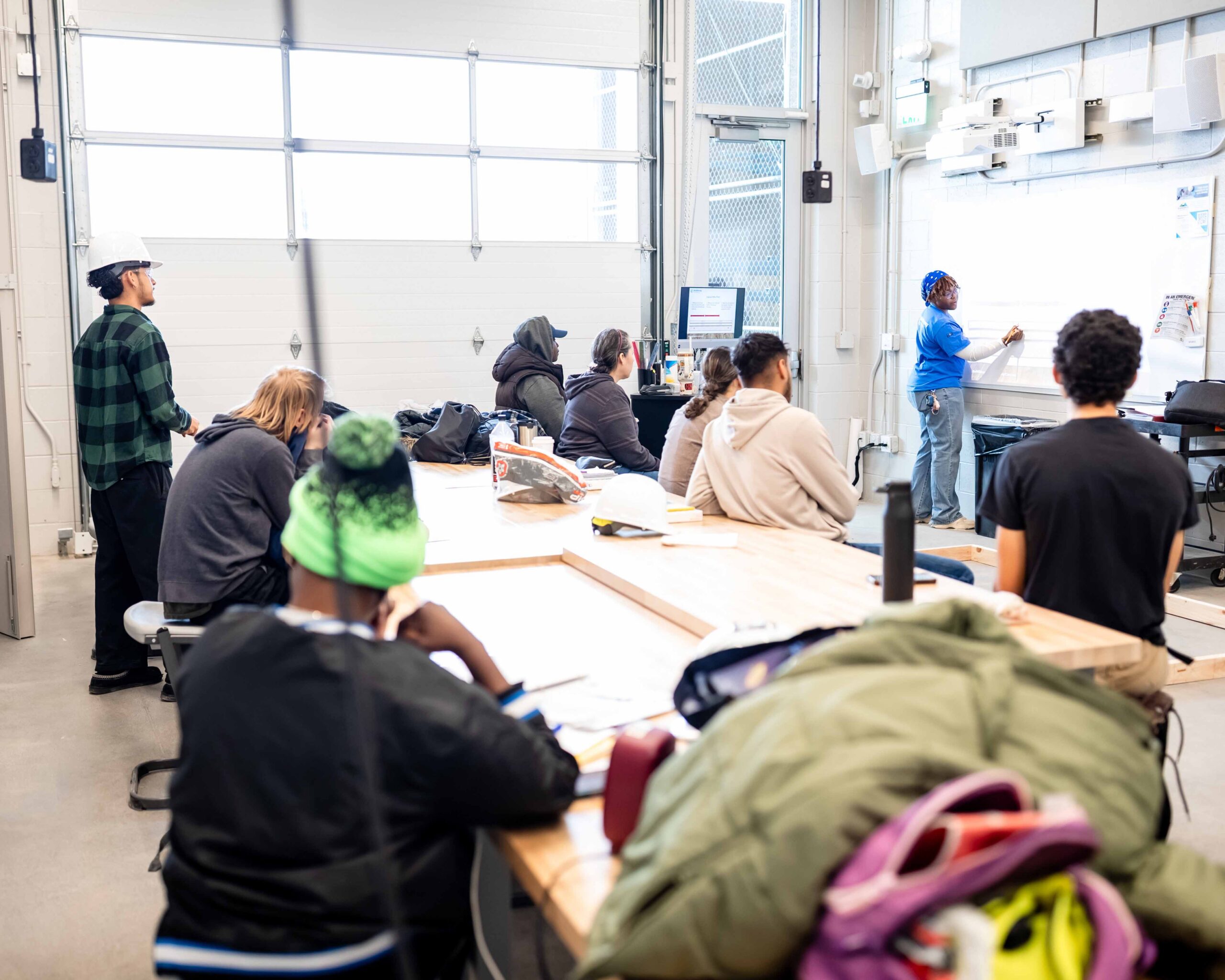 Students sit at workbenches in a workforce training classroom while an instructor writes on a whiteboard at the front of the room.