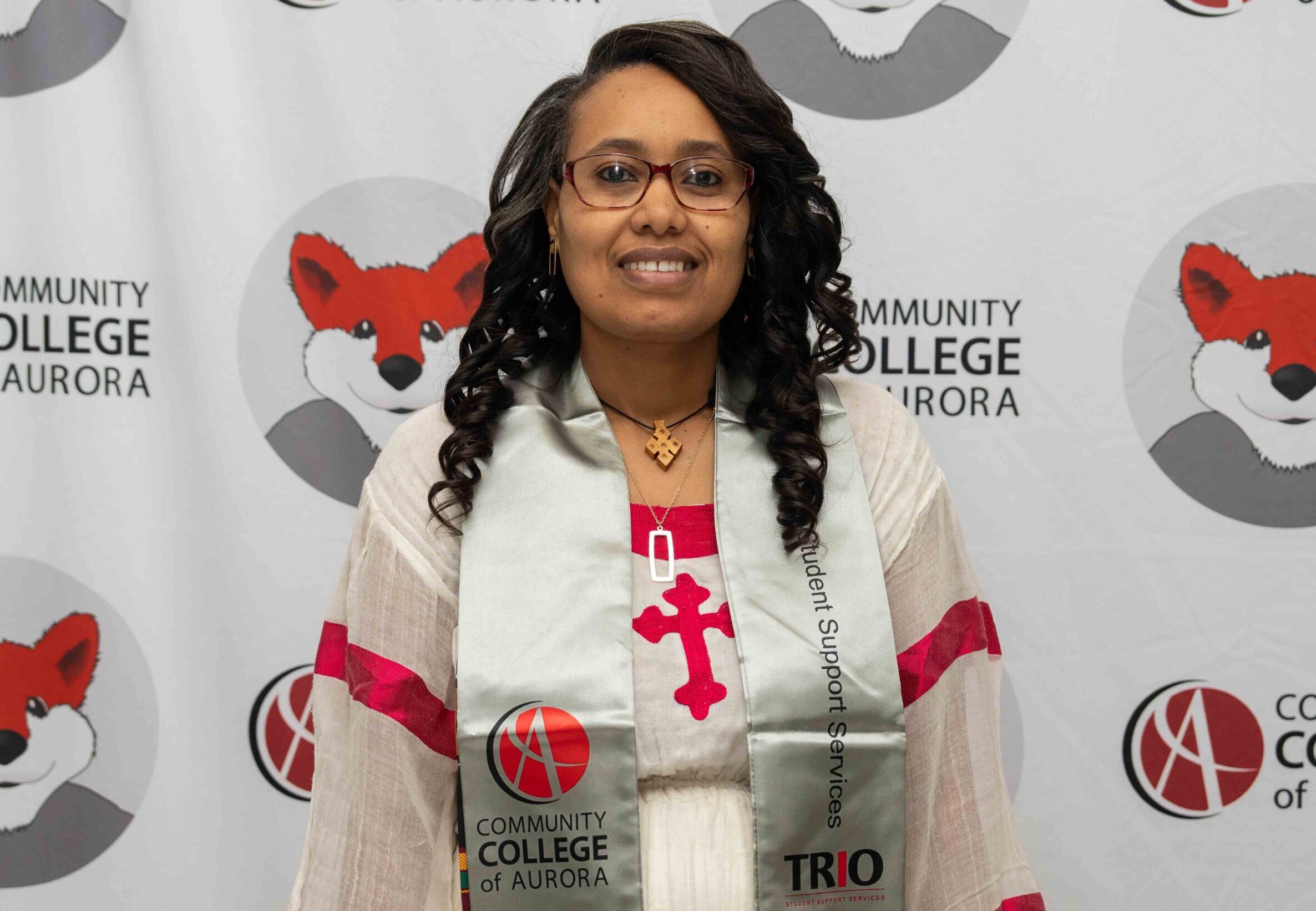 Photo of Abaynesh Berecha in ceremonial stole stands before a Community College of Aurora and fox-logo backdrop, conveying pride.