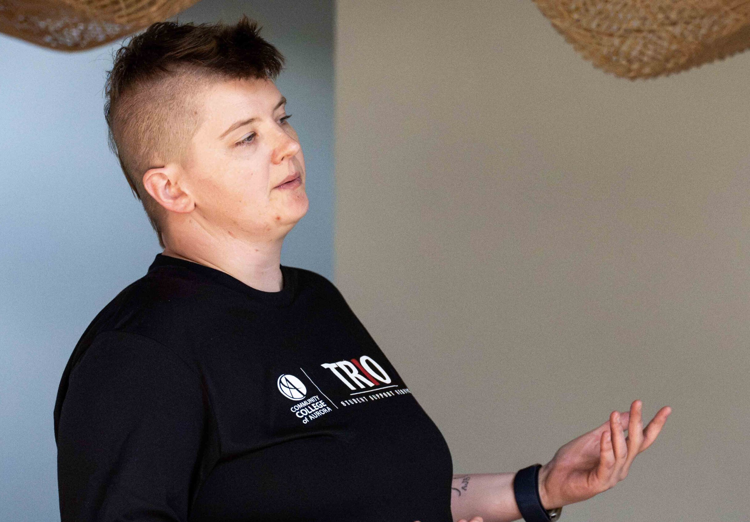 Photo of Abby Vestecka in a black TRIO shirt gesturing while speaking indoors, conveying confidence and engagement in a conversational setting.