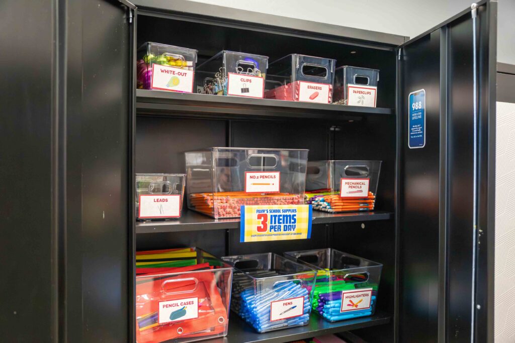 Open black storage cabinet with labeled bins of school supplies—pens, pencils, erasers, markers, and paper—neatly arranged, conveying order.