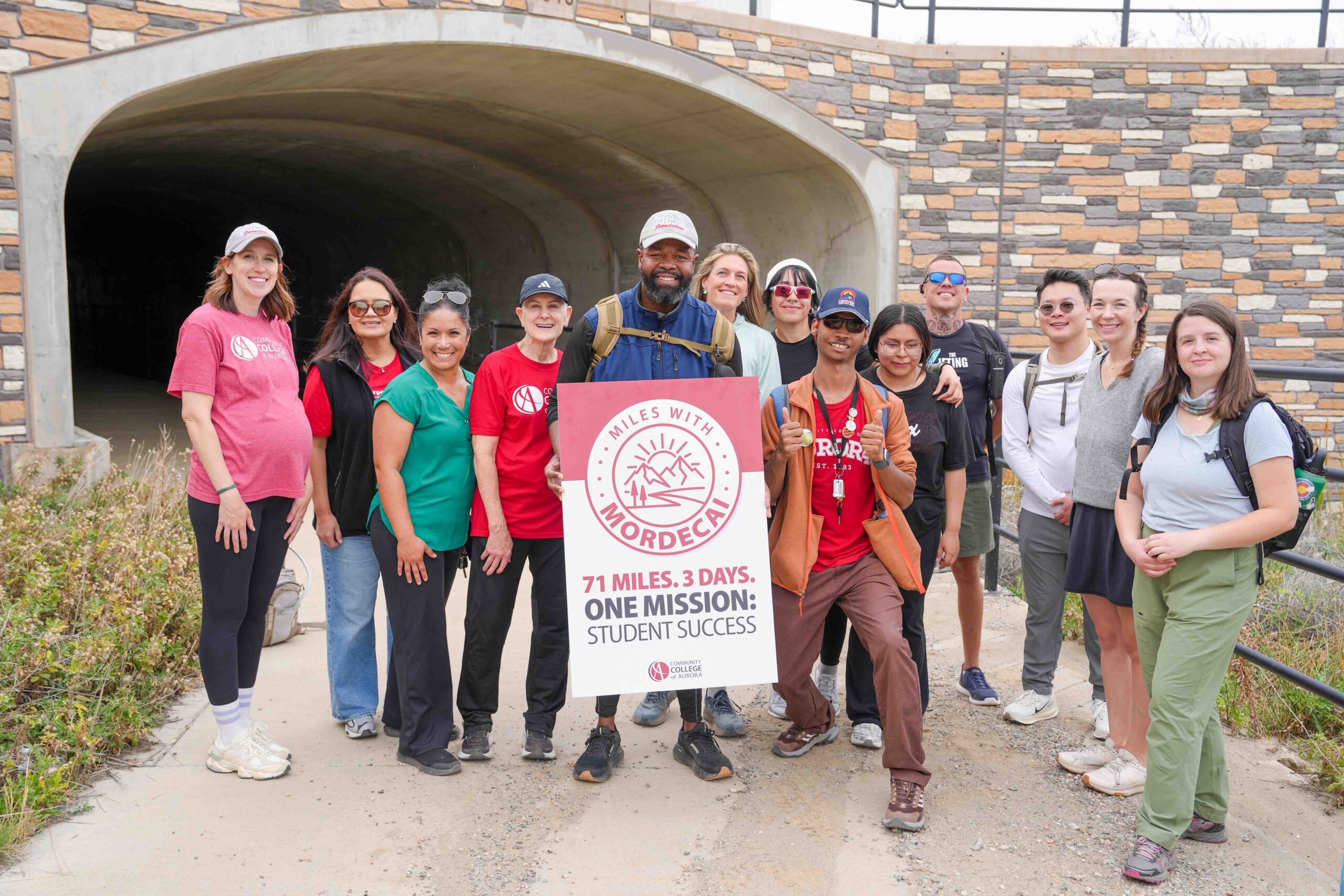Group of people standing in front of a tunnel entrance and holding a sign about MILES WITH MORDECAI. The sign also includes the Community College of Aurora logo. Participants gather on a trail during an outdoor walking event.