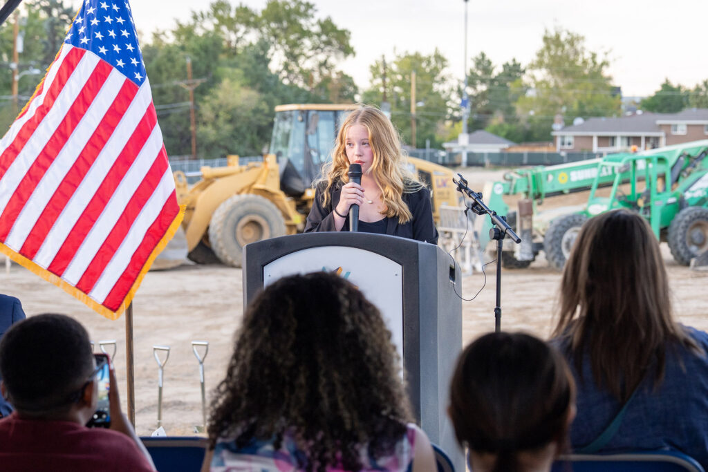 Hailey Jolly speaks into a microphone at an outdoor ceremony, with an American flag, construction equipment, and seated audience in view.