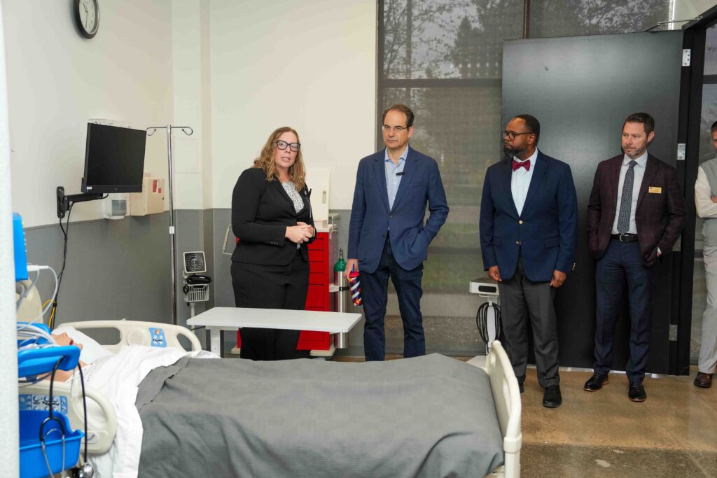 Phil Weiser listens as Jenn Dale speaks in a healthcare simulation lab. A hospital bed with a patient simulator, medical equipment, and several attendees are visible during the campus tour.