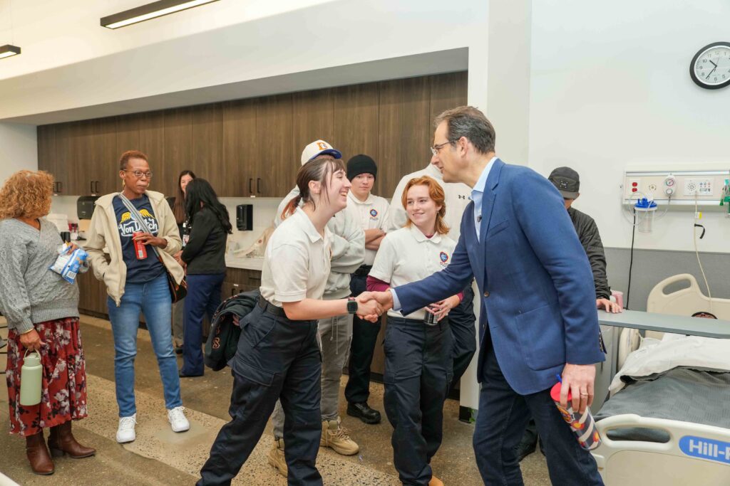Phil Weiser shakes hands with a student in a healthcare training classroom while other students and visitors look on. A hospital bed, medical equipment, and classroom cabinetry are visible in the background.