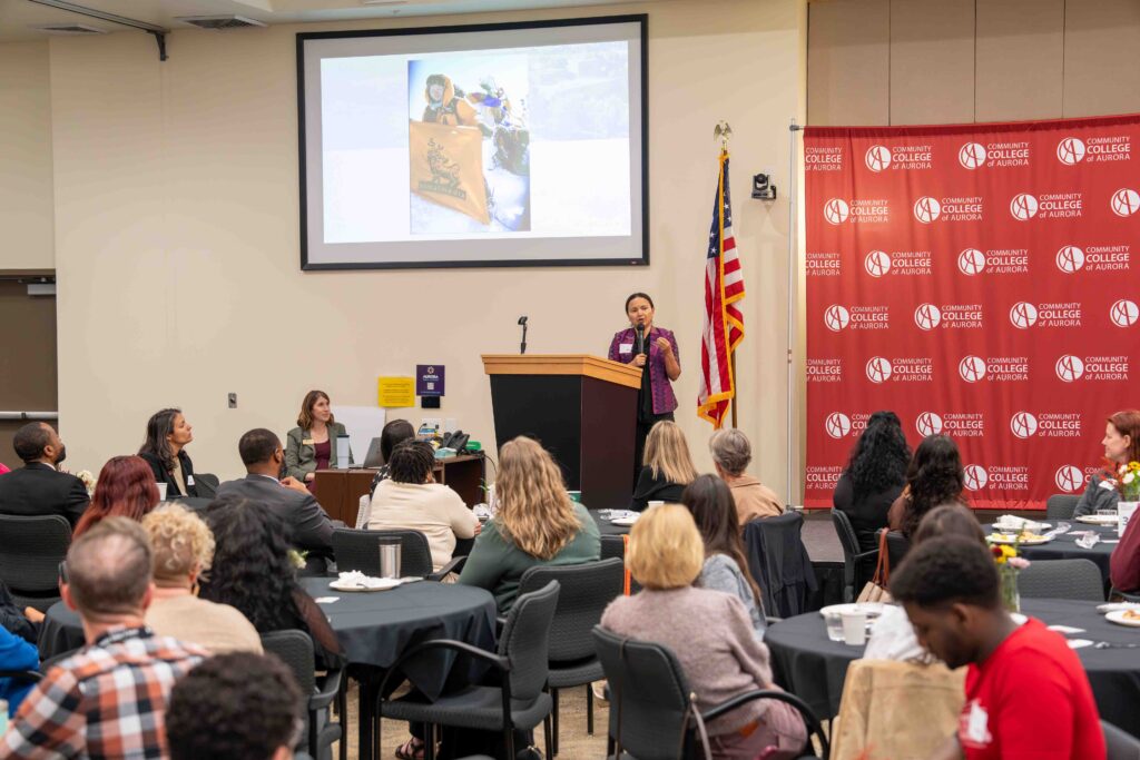 Shailee Basnet addresses a seated audience beside a podium, with a projected slide, U.S. flag, and red college banner creating a formal, attentive event atmosphere.